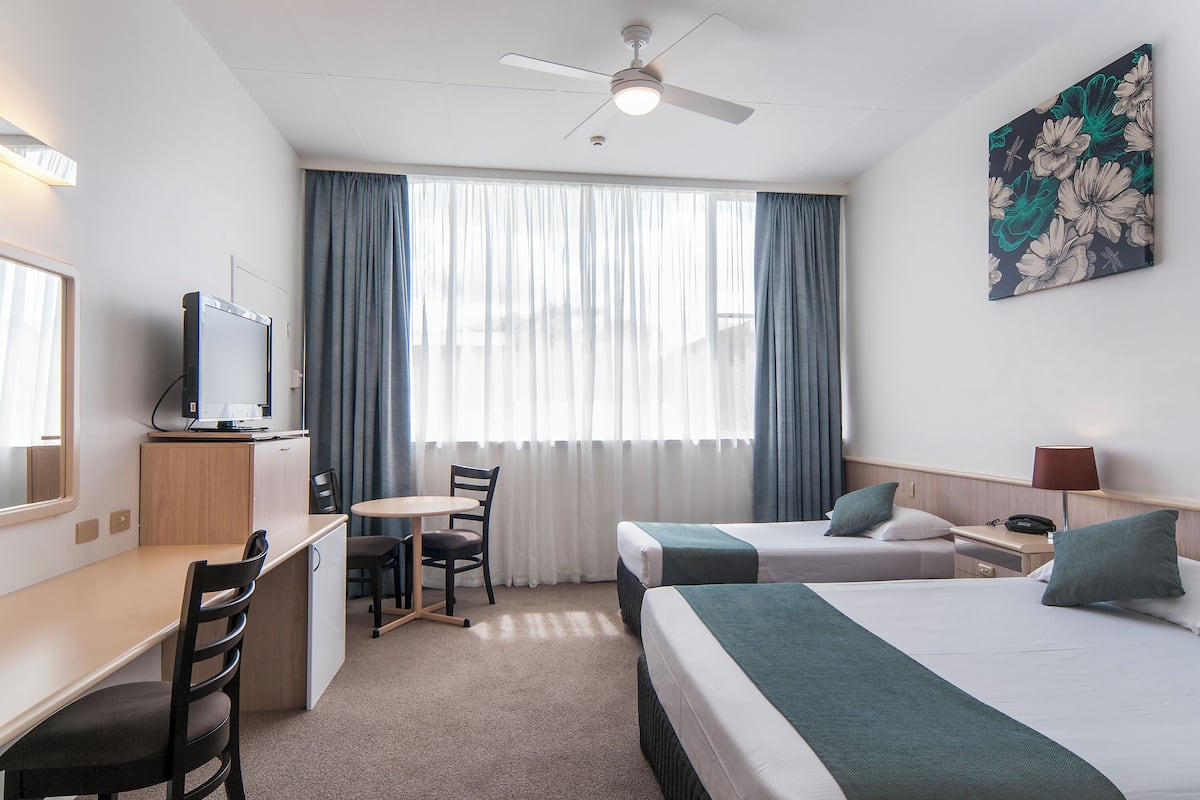 Hotellihuone, jossa on työpöytä, kaksi sänkyä, kattotuuletin, televisio ja taulu seinällä.