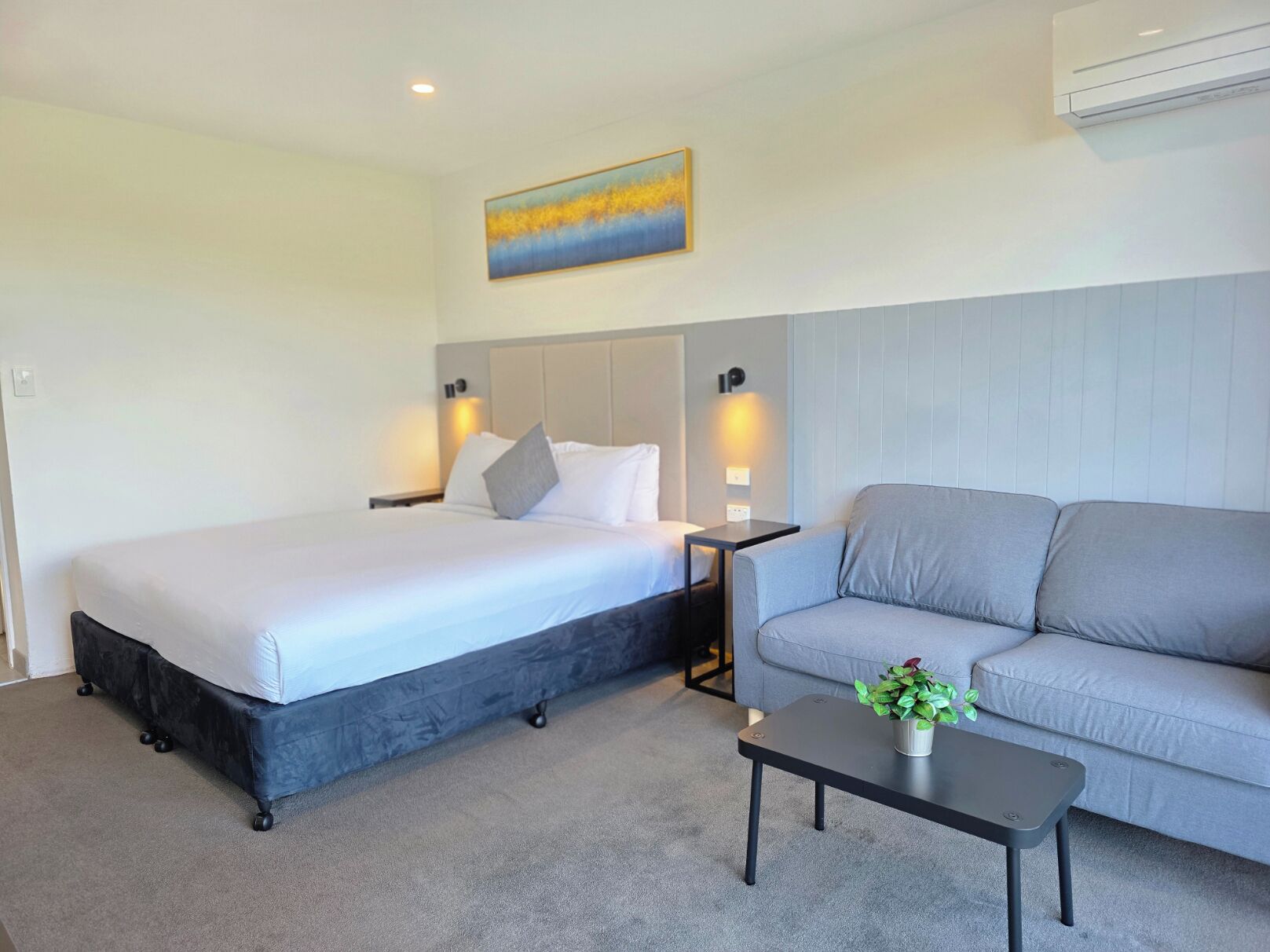 A hotel room with a bed, a grey sofa, a small table with a plant, and a painting on the wall.