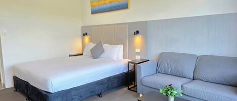 A hotel room with a bed, a grey sofa, a small table with a plant, and a painting on the wall.