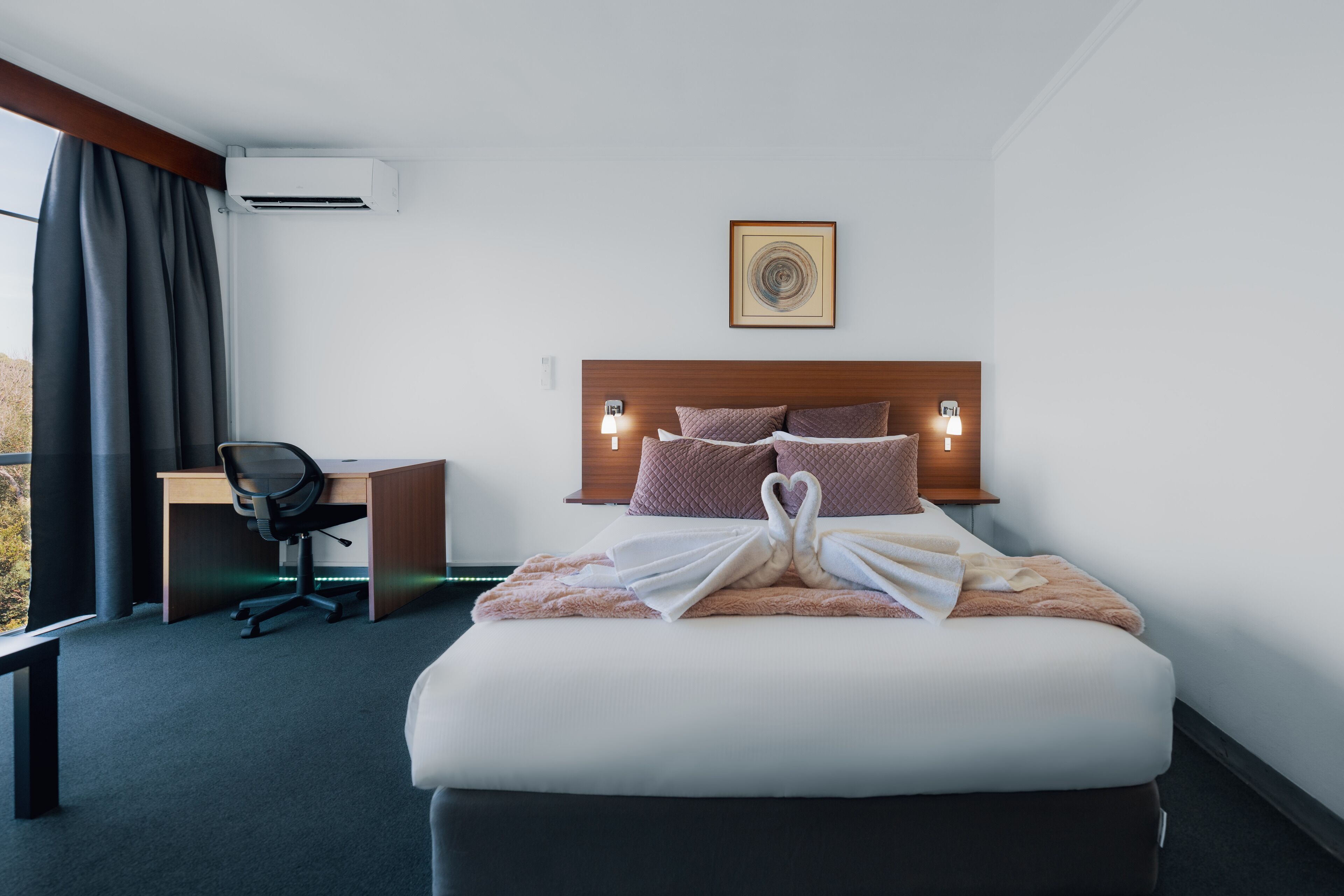A hotel room with a bed, a desk, a chair, a wall-mounted air conditioner, and a framed picture on the wall.