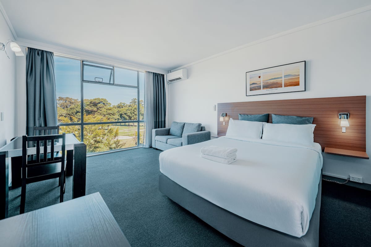 A modern hotel room with a large bed, a desk, and a sofa. There is a window with a view of trees and a cityscape.