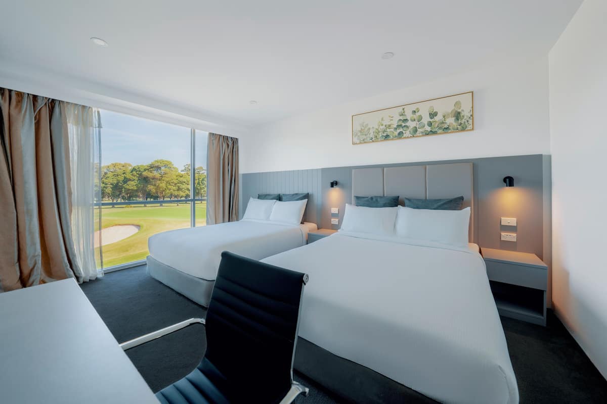 A hotel room with two beds, a desk, and a chair. There is a large window with a view of a golf course.