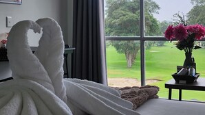 Une chambre d’hôtel avec une grande fenêtre, un lit recouvert de draps blancs, un vase rempli de fleurs et un tableau accroché au mur.