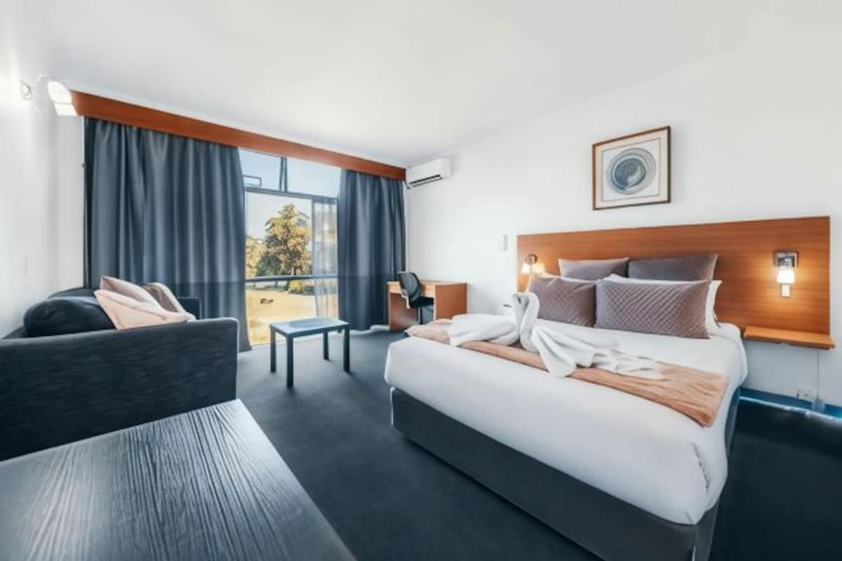 A modern hotel room with a large bed, a sofa, a small table, and a view of the outdoors.