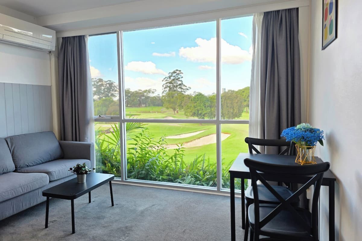 A room with a grey sofa, a small table, a chair, and a view of a golf course through large windows.