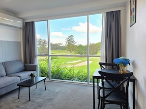 A room with a grey sofa, a small table, a chair, and a view of a golf course through large windows.