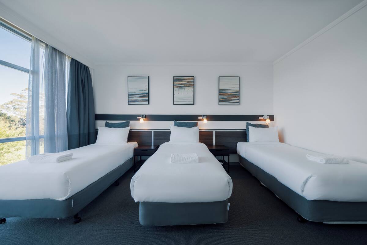 A hotel room with two single beds, a large window with curtains, and three wall-mounted artworks.