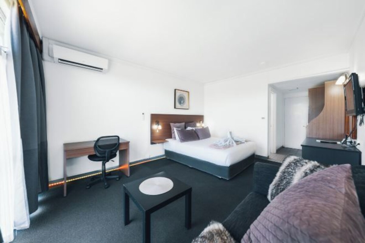 A modern hotel room with a bed, desk, chair, and a small table.