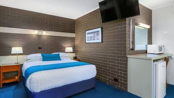 A hotel room with a large bed, a flat-screen TV mounted on the wall, a small refrigerator, and a bedside table with a lamp.