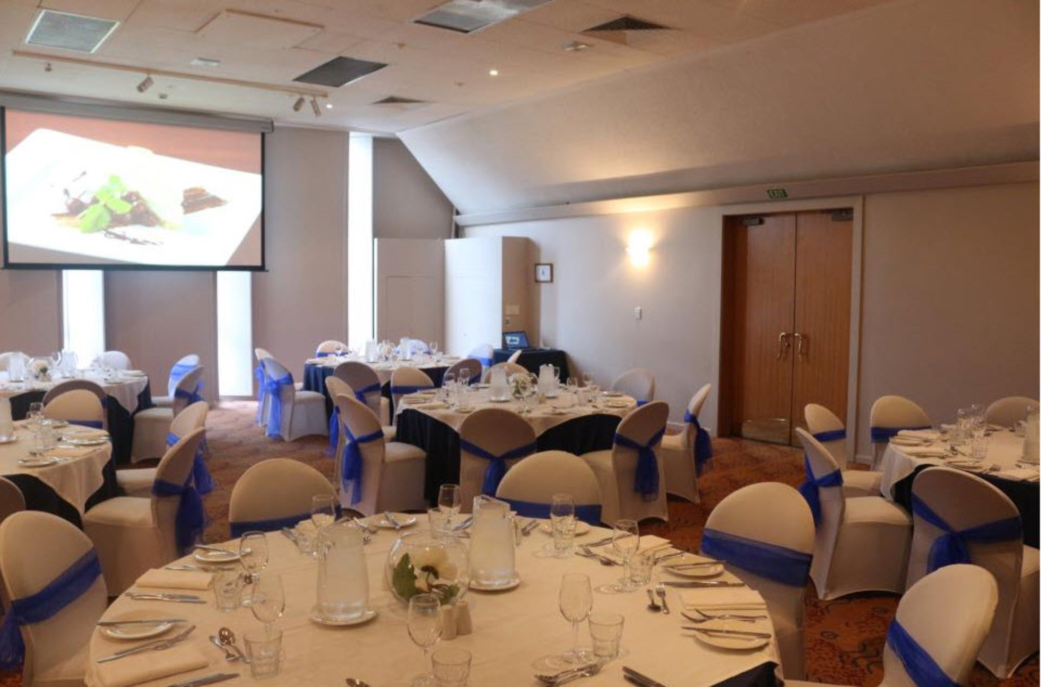 A banquet hall with round tables set for dining, each with white tablecloths and blue sashes.
