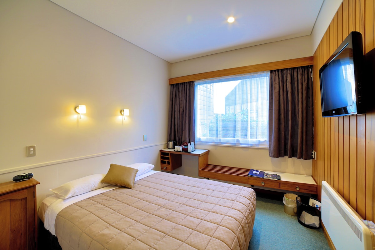 Habitación de hotel con cama, mesita de noche, lámpara, televisor, ventana con cortinas y vistas al exterior.