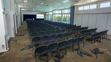 Un salón de conferencias con filas de sillas, una pantalla de proyección y ventanas amplias.