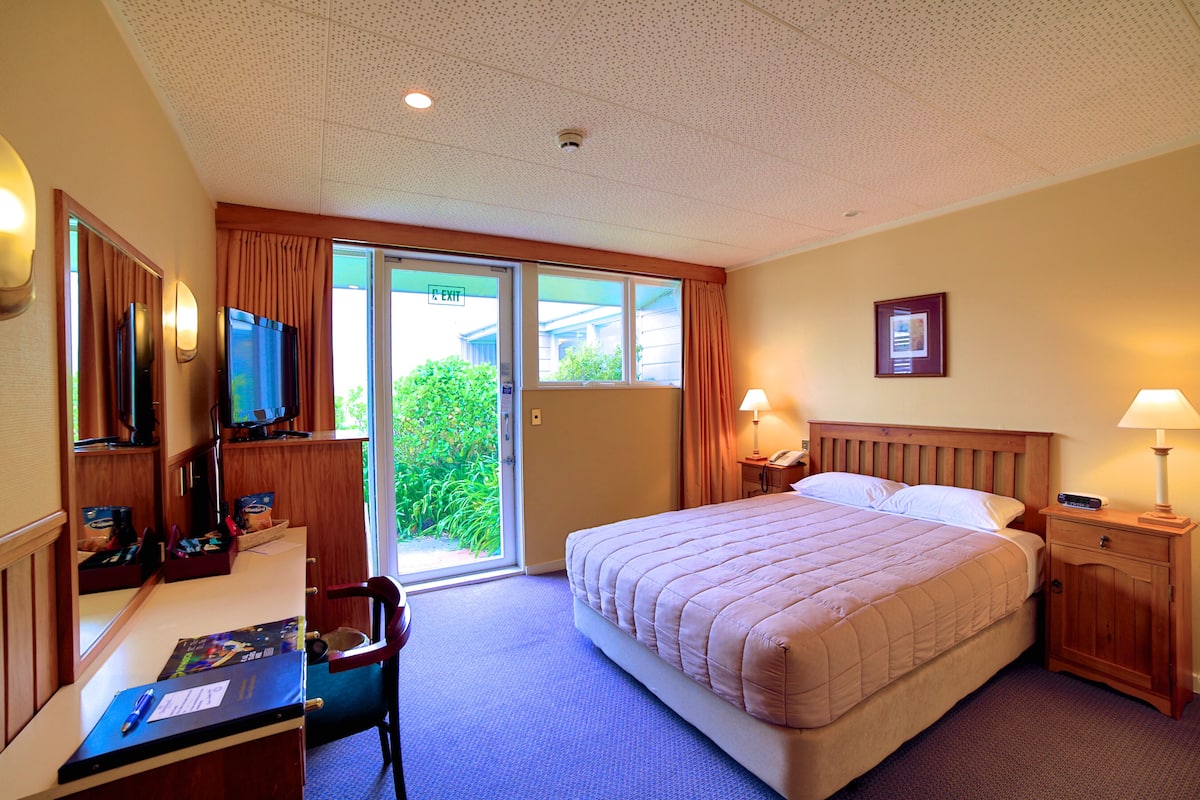 Une chambre d’hôtel avec un grand lit, un bureau avec un ordinateur, une télévision et une porte-fenêtre coulissante donnant sur l’extérieur.