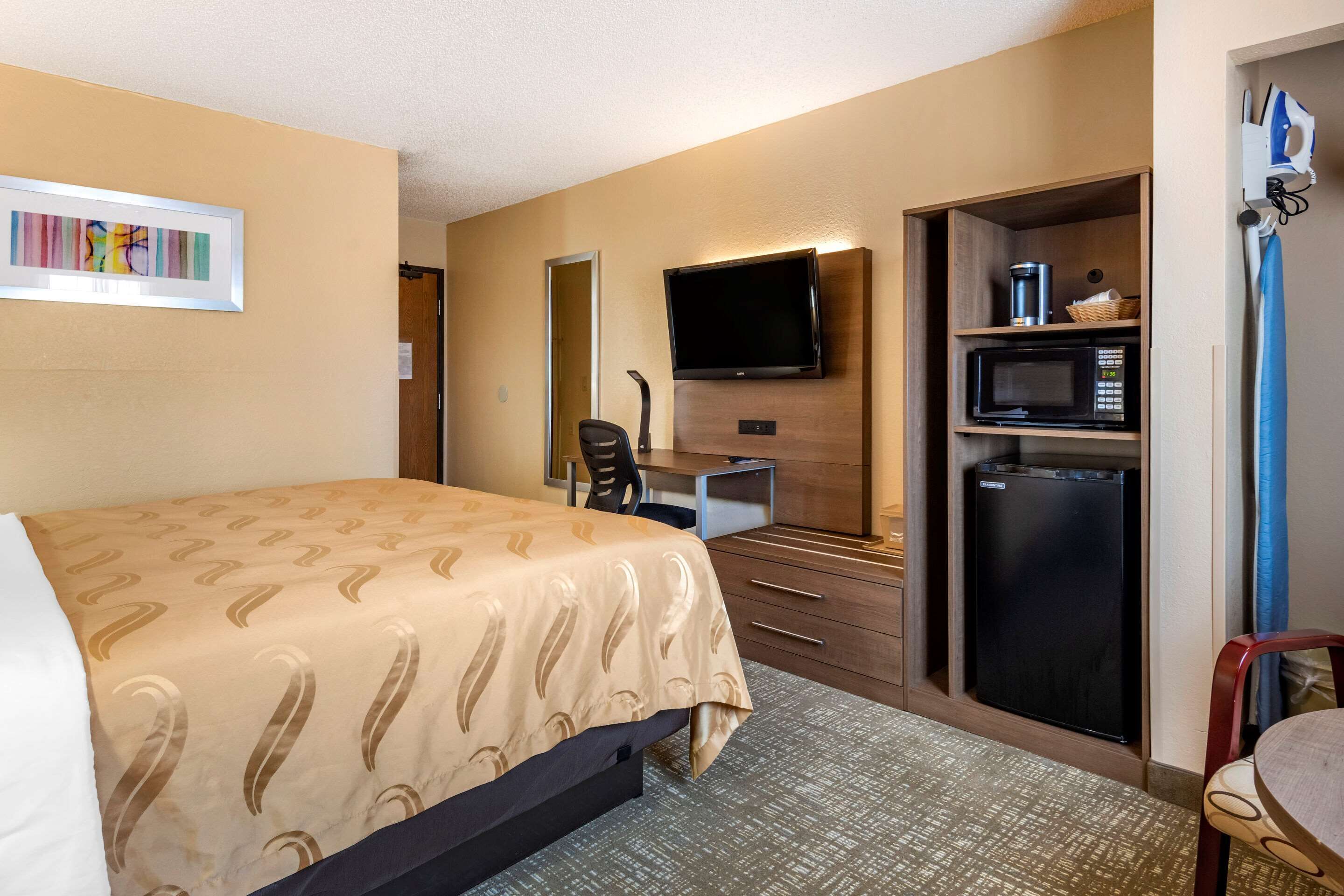 A hotel room with a bed, a desk with a computer, a television, a microwave, and a refrigerator.