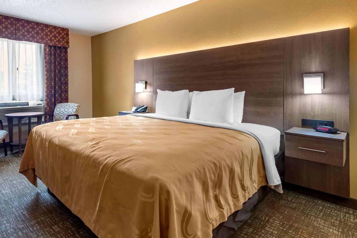 Ein Hotelzimmer mit einem großen Bett, einem Nachttisch mit Telefon und digitaler Uhr, einem kleinen Schreibtisch mit Stuhl sowie einem Fenster mit Vorhängen.