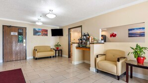 Un hall de hotel con dos sillones color beige, una estación de café, un televisor montado en la pared y una planta en maceta roja.