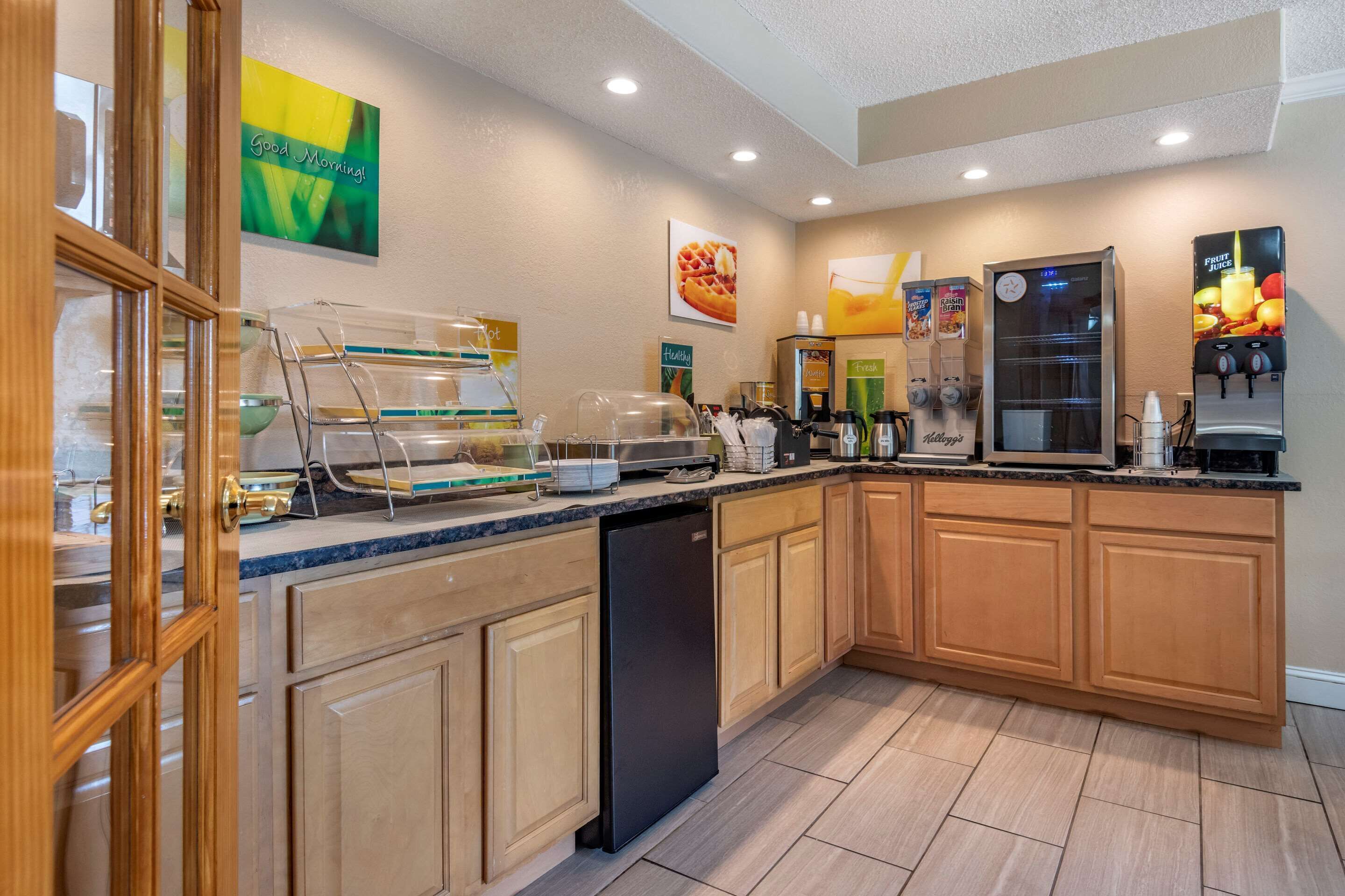 A hotel kitchen with a self-service breakfast station, a coffee machine, a refrigerator, and a fruit juice dispenser.