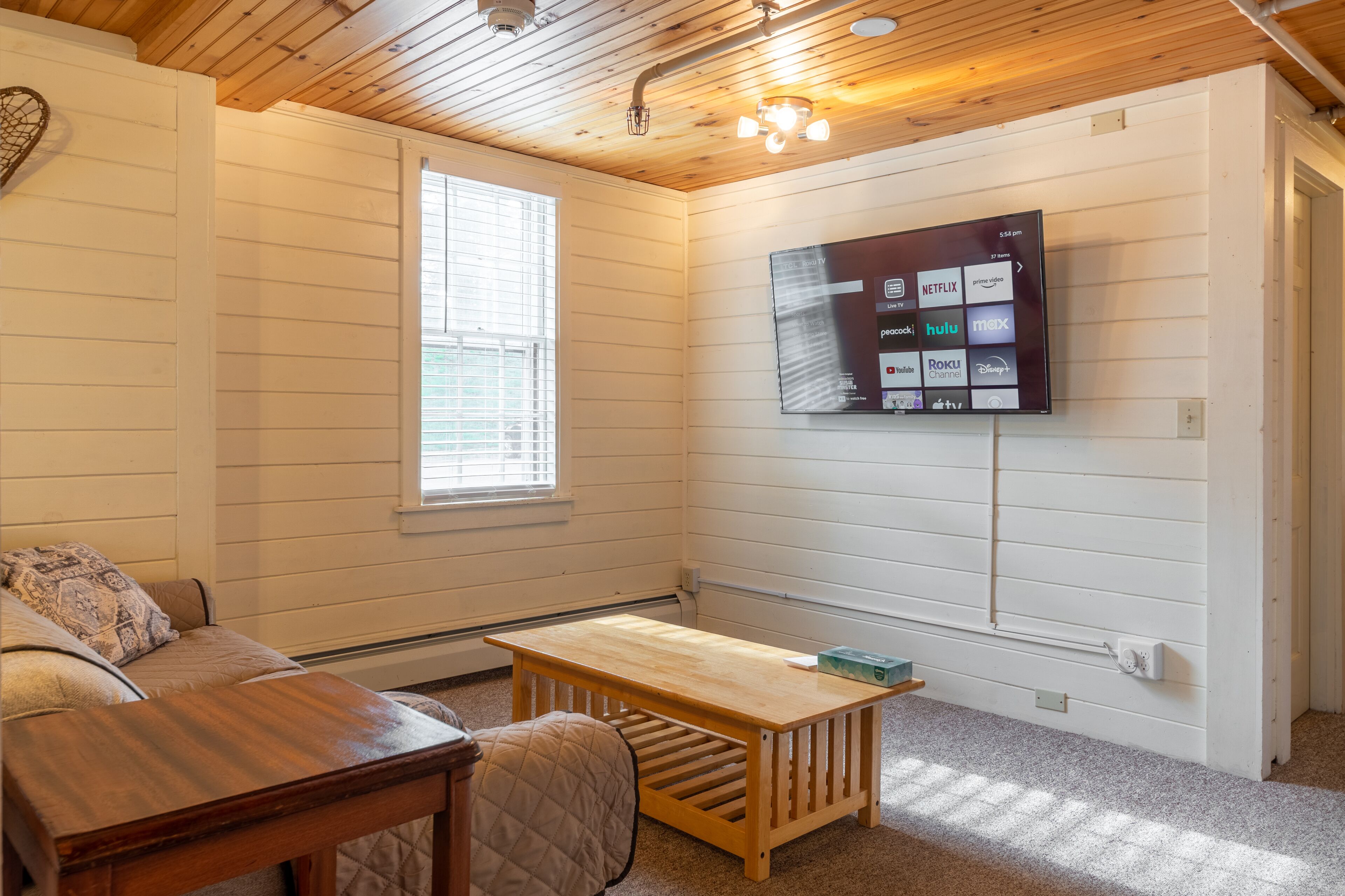 A cozy room with a wooden ceiling, a TV mounted on the wall displaying various streaming services, a small wooden table, and a cushioned bench.
