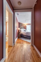 Un pasillo estrecho que conduce a un dormitorio con cama, mesita de noche, lámpara y cortinas.