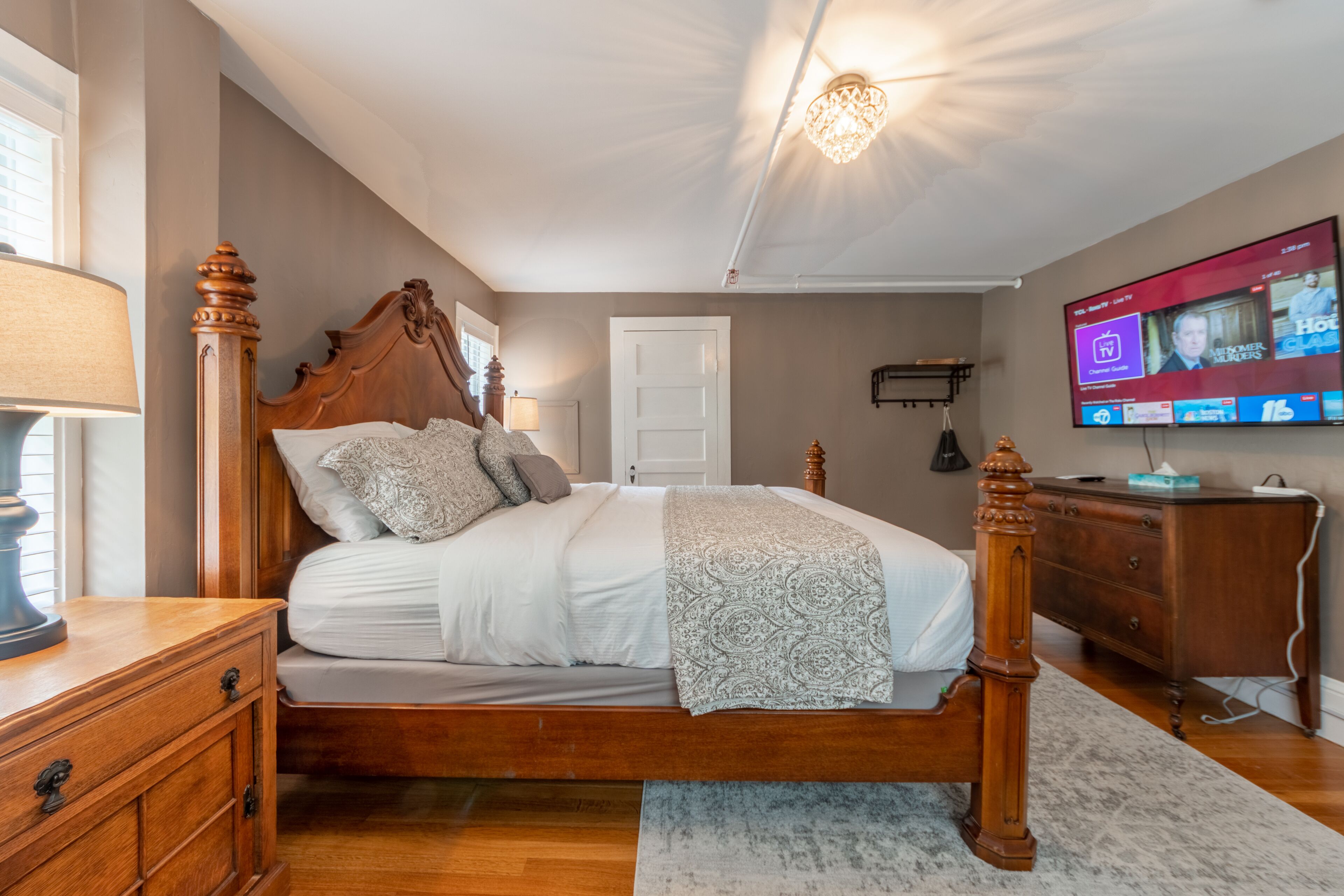 Un dormitorio con cama de dos columnas, cómoda de madera, televisor montado en la pared y candelabro.