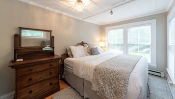 Quarto com cômoda de madeira, cama com roupa de cama estampada, lustre e janela com persianas.