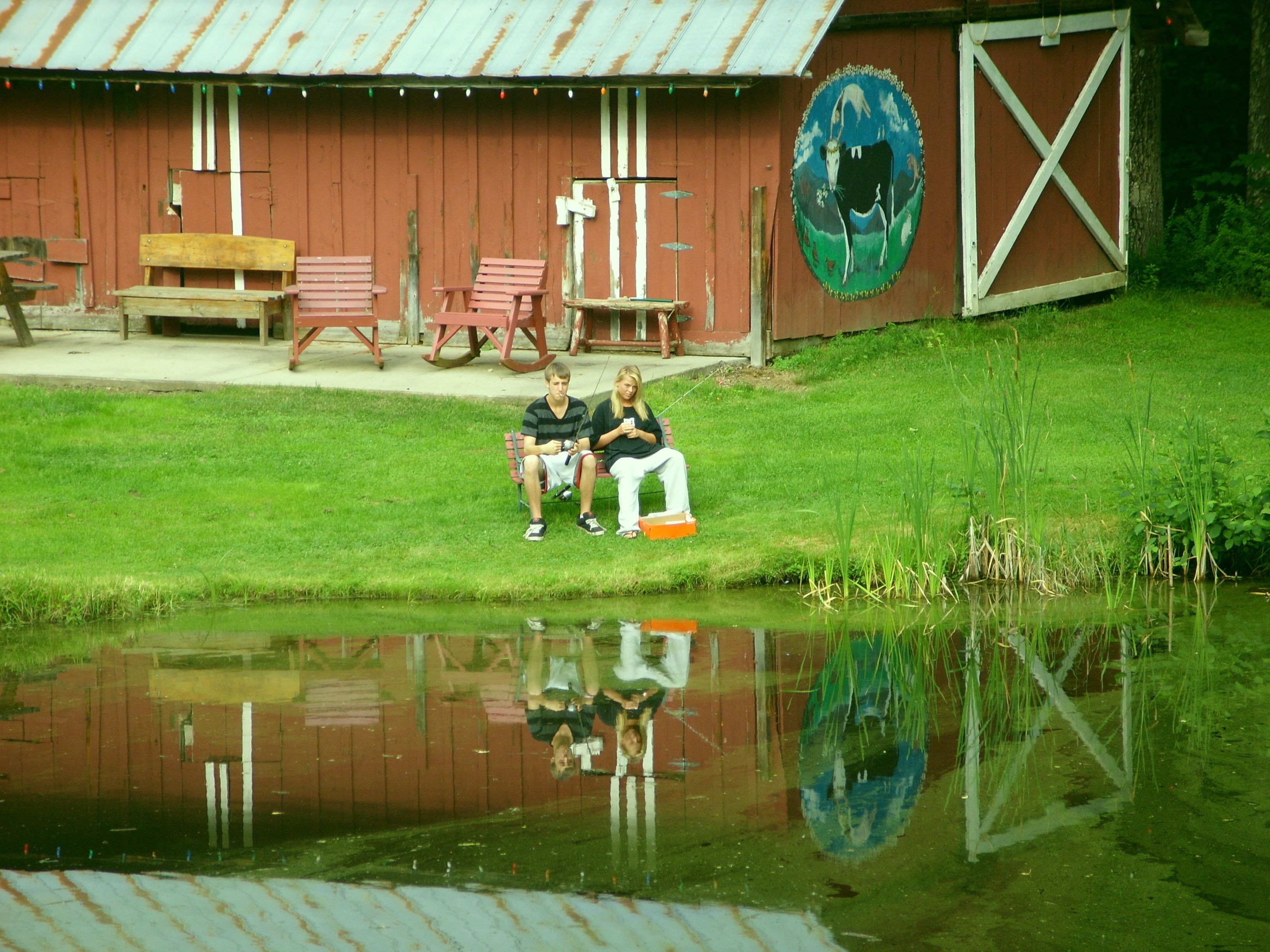 Two people sitting on a bench by a pond with a barn and picnic tables in the background.