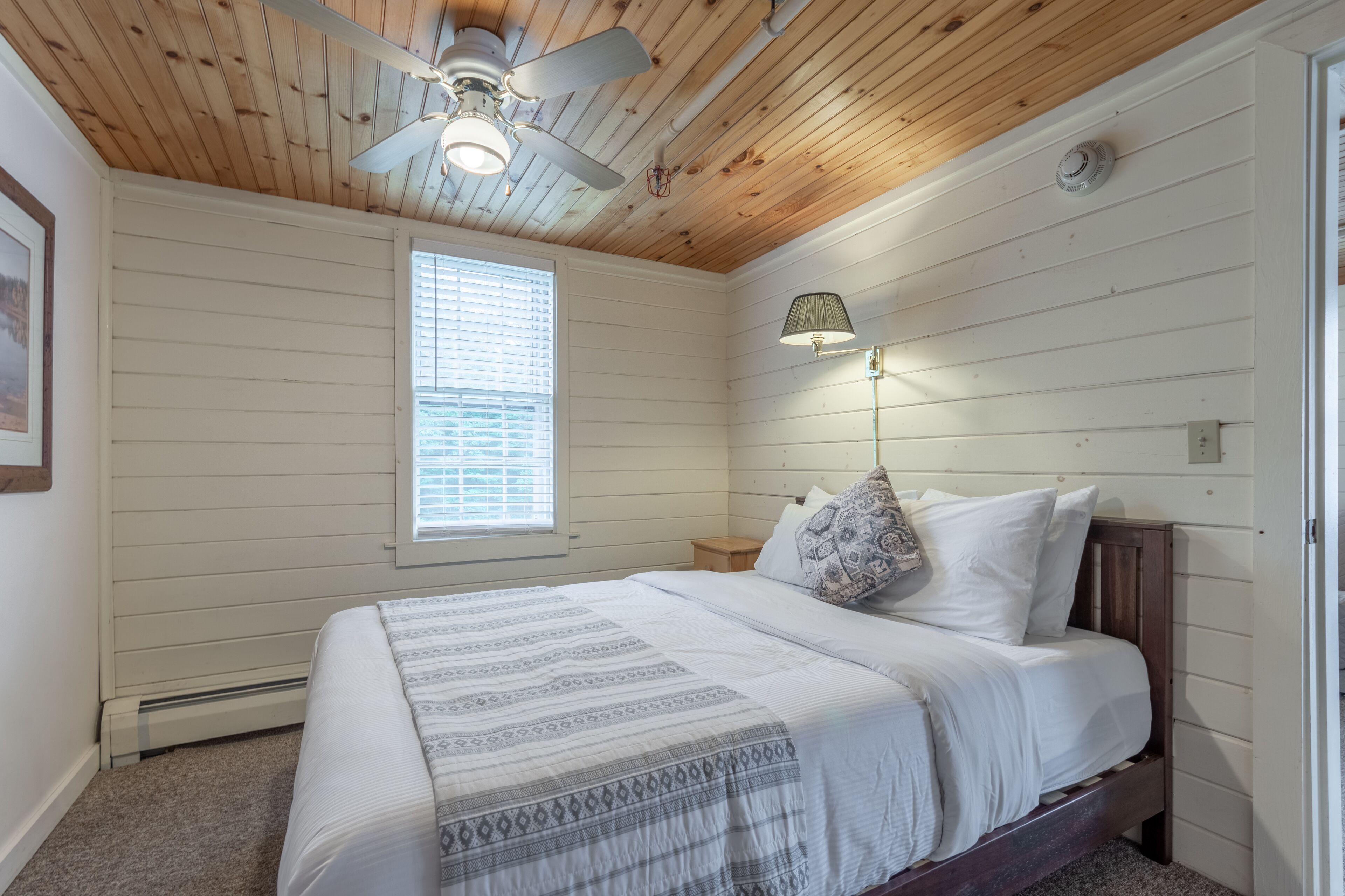A bedroom with a wooden ceiling, a bed with white bedding, a window with blinds, and a wall-mounted lamp.