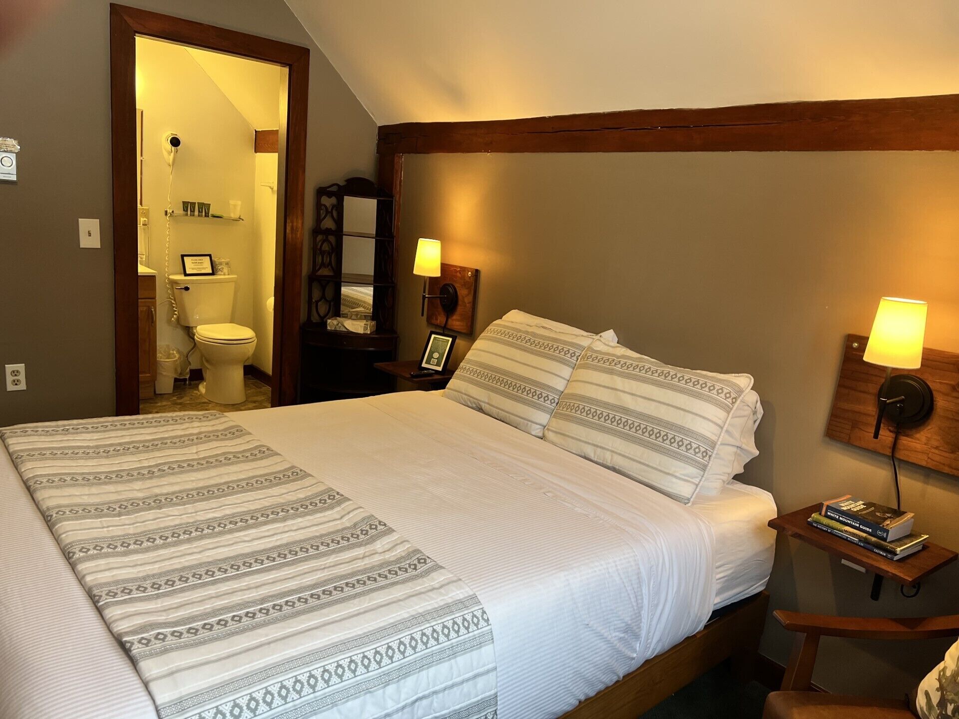 A hotel room with a large bed, two bedside lamps, a nightstand with books, a wooden headboard, and a bathroom visible through an open door.