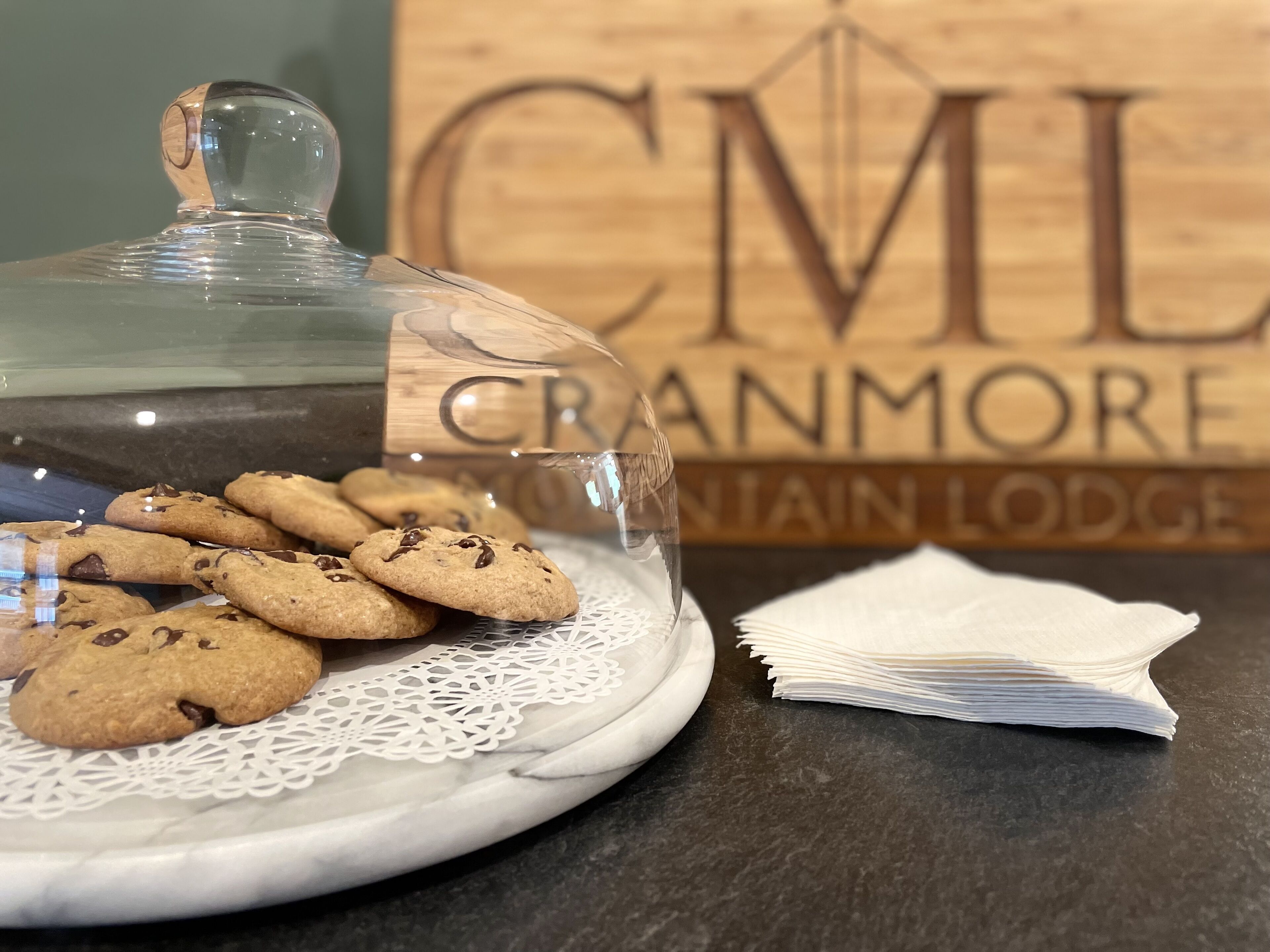 A tray of cookies with a glass dome cover, a lace doily, and a stack of napkins on a marble surface. In the background, there's a wooden sign with the text "CML Cranmore" and "Cranmore Lodge".