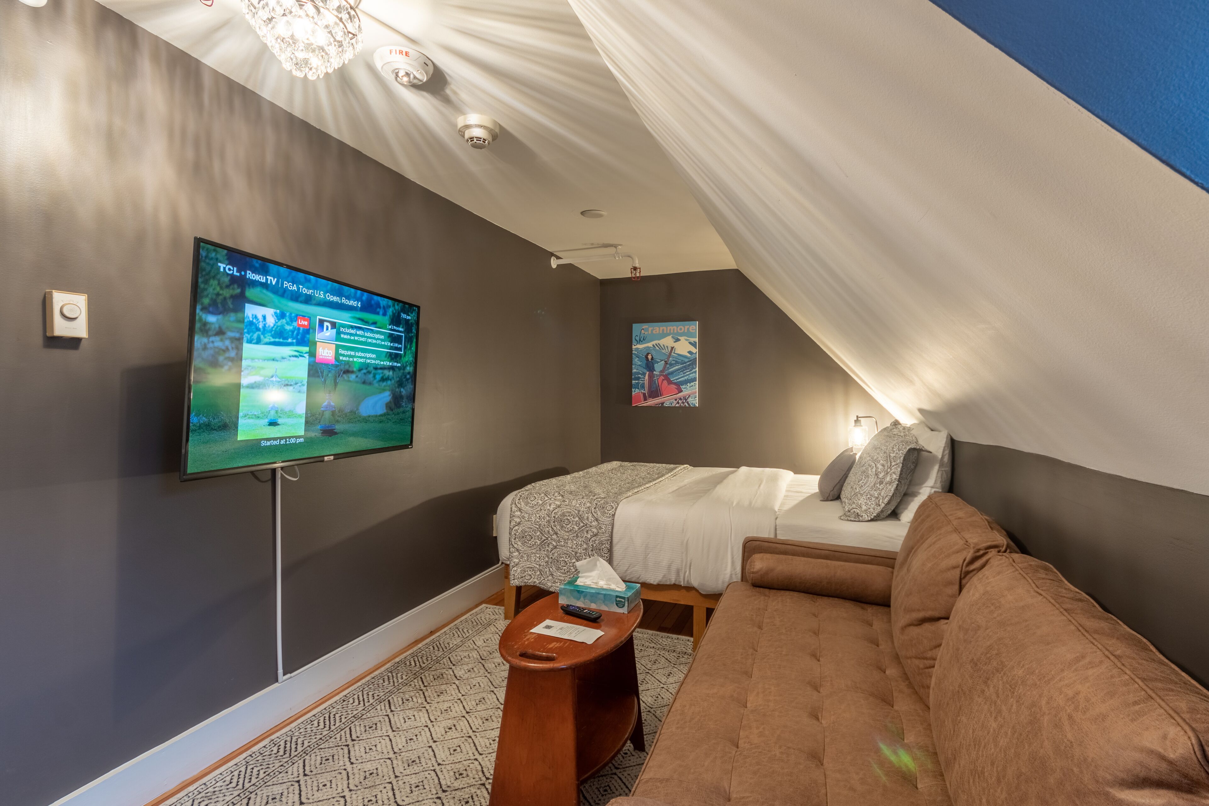 Une pièce confortable avec un grand téléviseur à écran plat fixé au mur, un lit recouvert d’une couverture blanche à motifs, un canapé marron et une petite table d’appoint en bois.