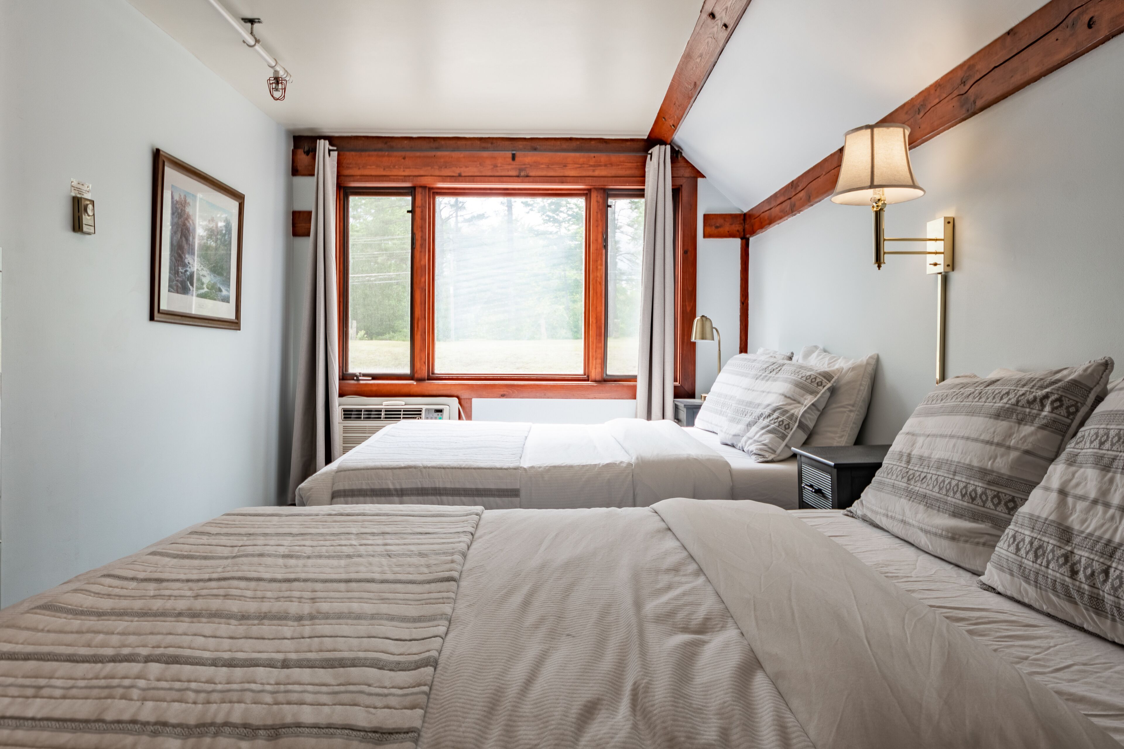 A bedroom with two beds, a large window, wooden beams, a bedside lamp, and a framed picture on the wall.