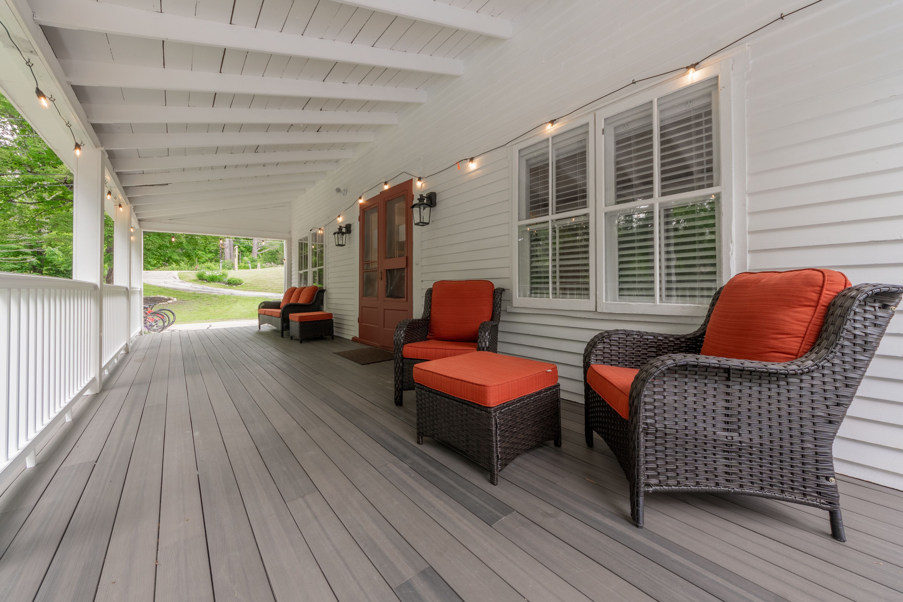 A covered porch with wicker furniture and orange cushions, white railings, and string lights.
