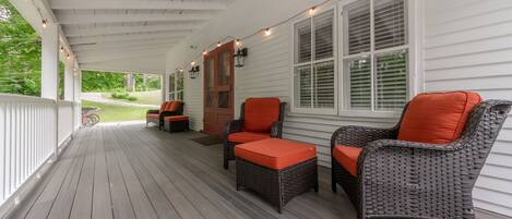 A covered porch with wicker furniture and orange cushions, white railings, and string lights.