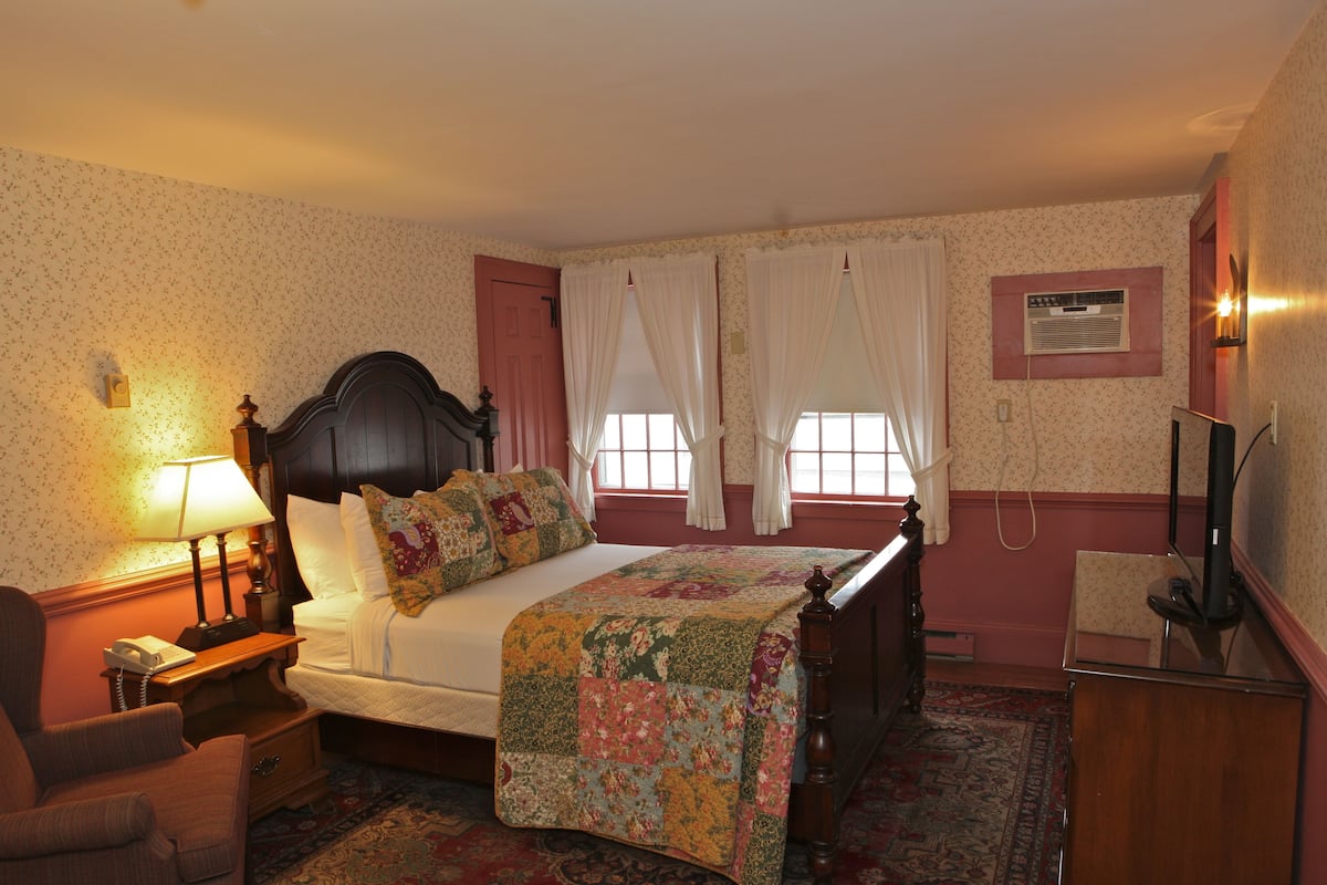 A hotel room with a bed, a nightstand with a lamp, a chair, a desk with a telephone, and a television.