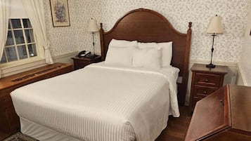 A neatly arranged bedroom with a large bed, a bedside table with a lamp, a nightstand, a wooden dresser, and a patterned rug.