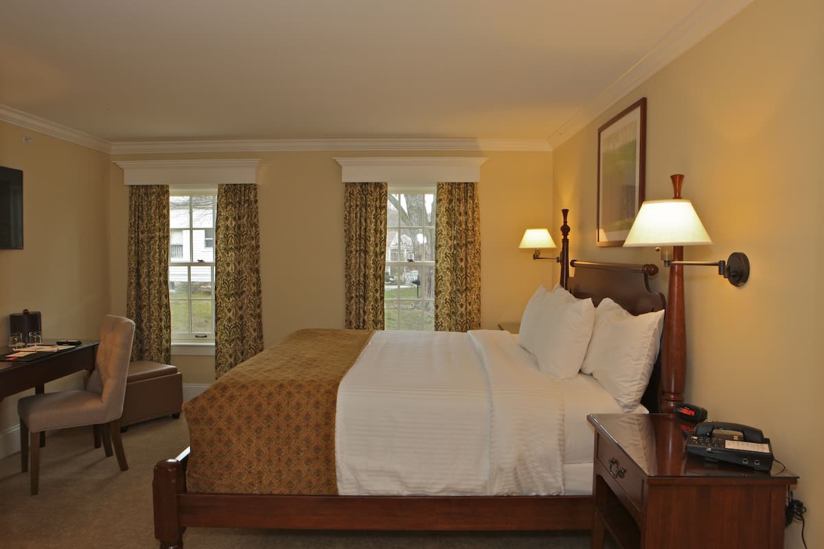 A hotel room with a large bed, a desk, a chair, and two windows with curtains.