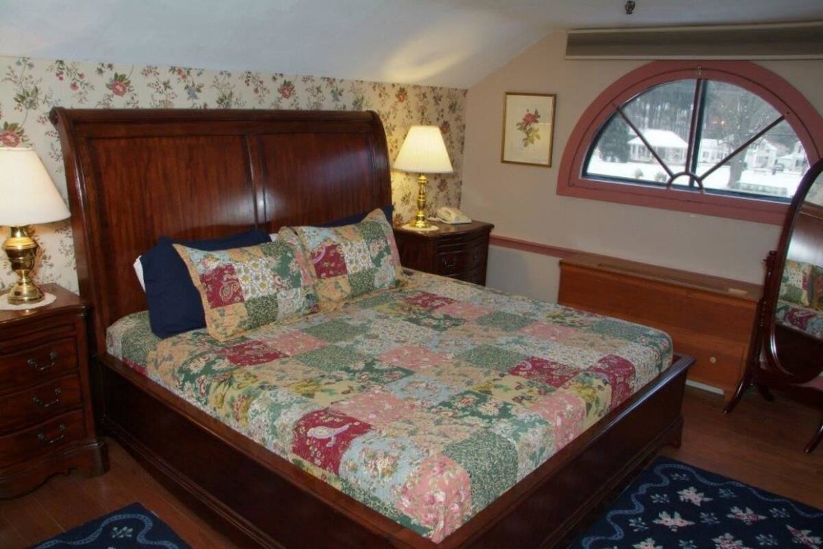 A bedroom with a wooden bed, floral wallpaper, a window with a view, and a nightstand with a lamp.