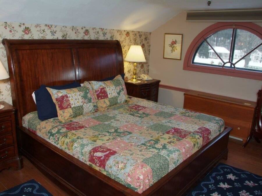 Ein Schlafzimmer mit einem hölzernen Bett, blumigem Wandpapier, einem Fenster mit Ausblick und einem Nachttisch mit Lampe.