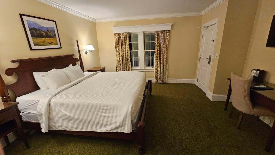 A hotel room with a large bed, a desk, a chair, a window with curtains, and a painting on the wall.