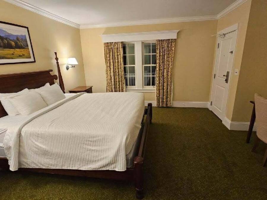 A hotel room with a large bed, a desk, a chair, a window with curtains, and a painting on the wall.