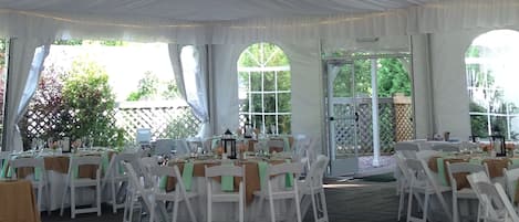 Un espacio para eventos con carpas, mesas y sillas blancas, candelabros y luz natural proveniente de ventanas.