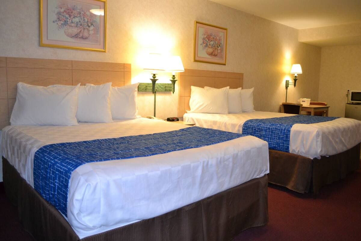 A hotel room with two beds, each with white linens and blue accents, a small desk with a chair, and two wall-mounted lamps.