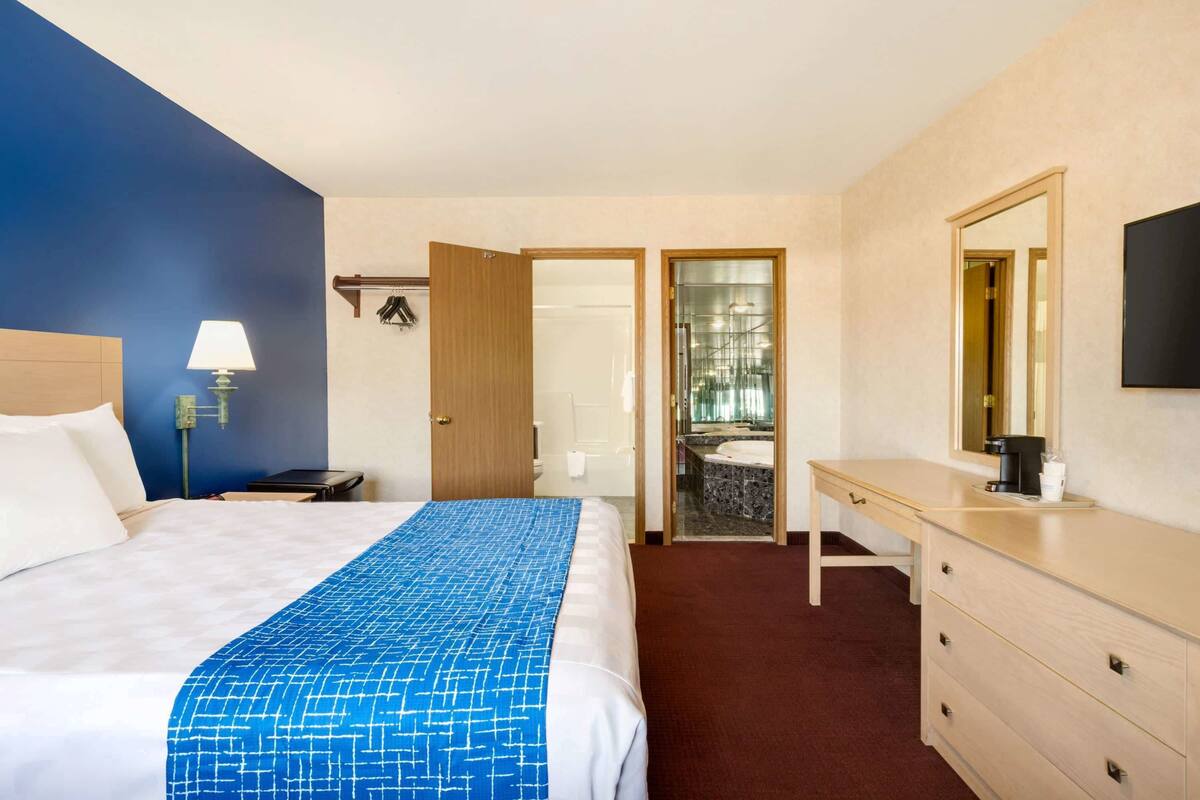 A hotel room with a bed, a desk, a chair, a lamp, a mirror, and a bathroom visible through an open door.