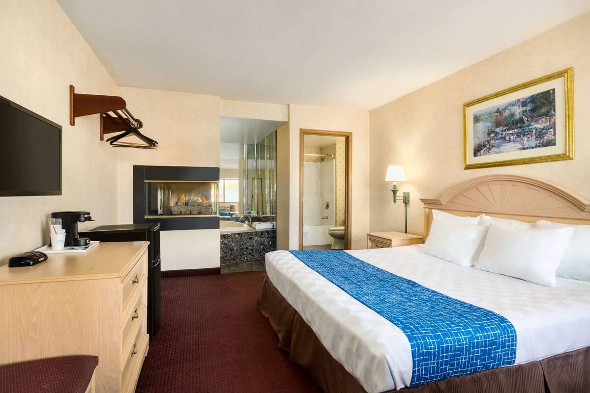 A hotel room with a bed, a desk, a dresser, a mirror, a painting, and a view of the bathroom.