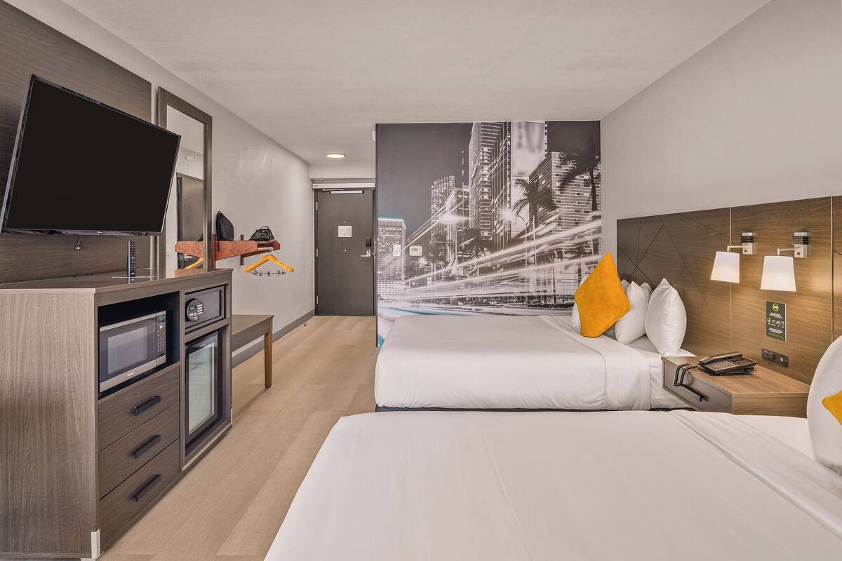 A hotel room with a bed, a TV, a desk with a microwave, and a cityscape wall mural.