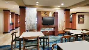 Un vestíbulo de hotel con un televisor de pantalla plana montado en una pared, rodeado de pilares de madera y una alfombra estampada. Mesas y sillas están dispuestas para comer.