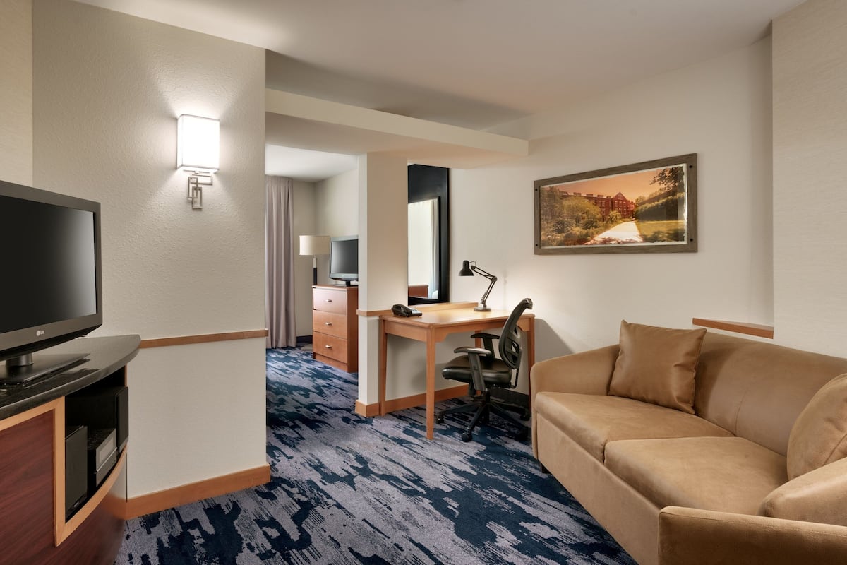 A hotel room with a flat-screen TV, a desk with a chair, a sofa, a painting on the wall, and a patterned carpet.