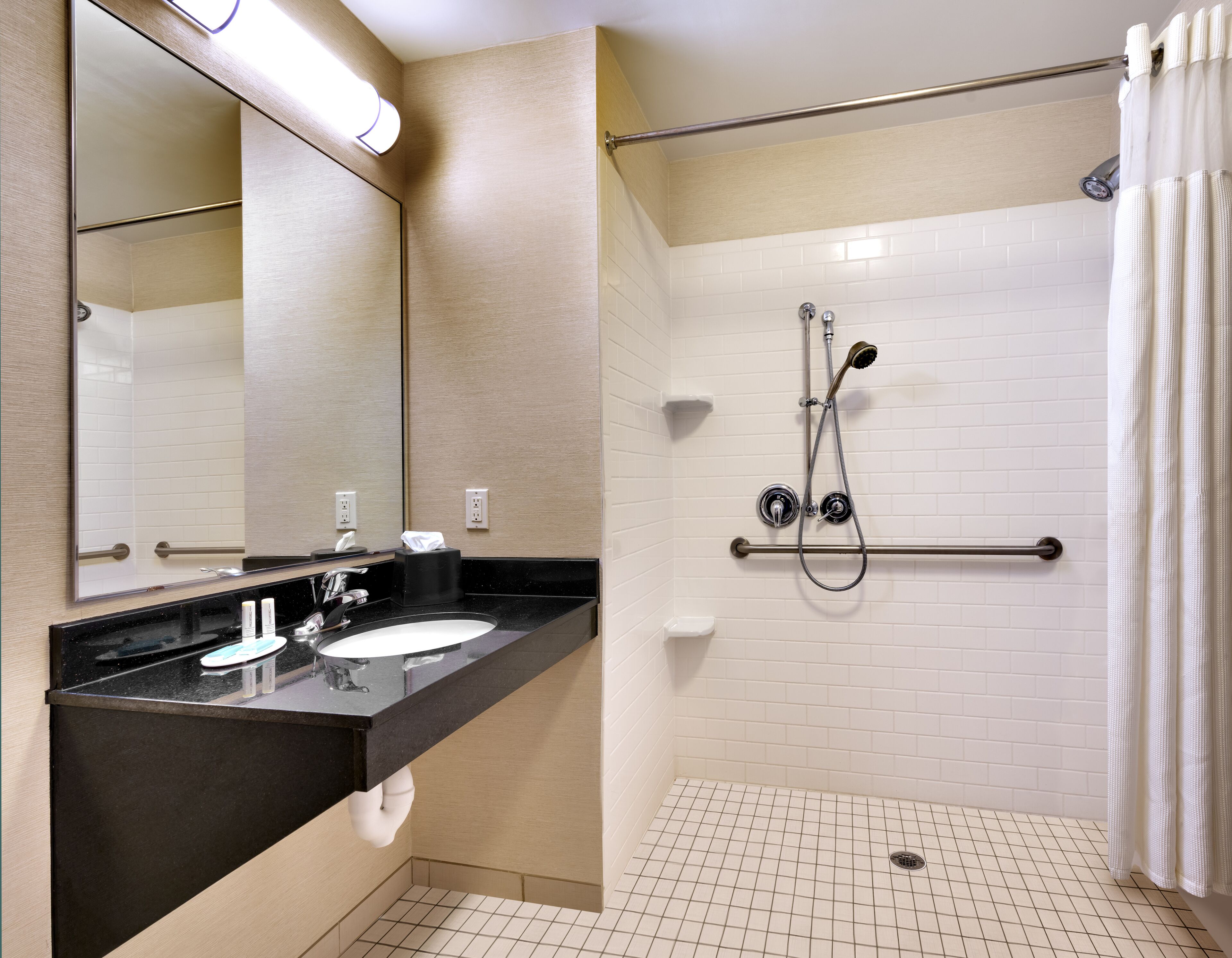 A bathroom with a black countertop sink, a mirror, a towel dispenser, and a shower with a grab bar.