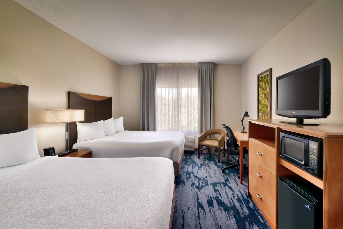 A hotel room with two beds, a desk with a flat-screen TV, a microwave, and a chair.