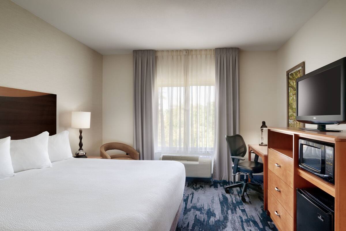 A hotel room with a large bed, a desk with a computer, a television, and a window with curtains.